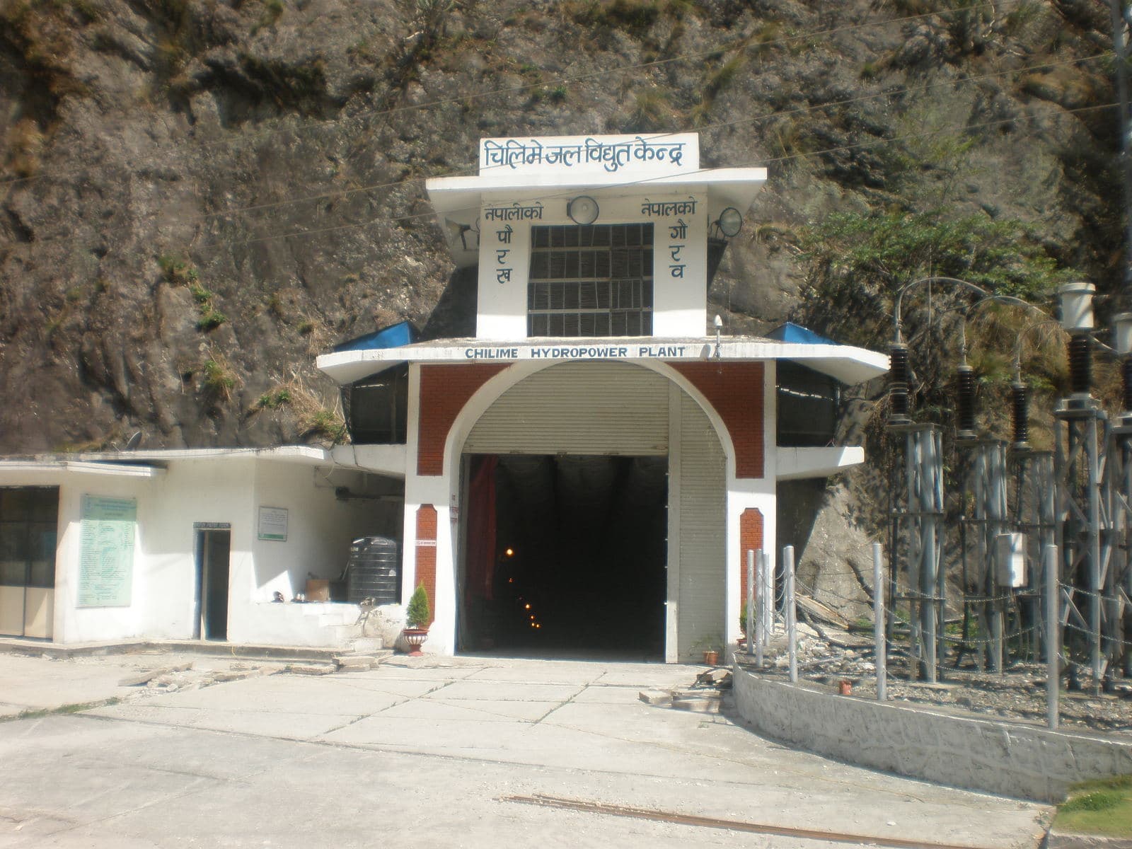 The arched tunnel entrance to the Chilime hydropower plant in Nepal, built into a steep rock face, with lights visible inside the dark tunnel and switchyard equipment to the right.