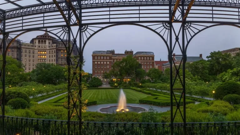 The Conservatory Garden in Central Park, New York, with manicured lawns, symmetrical hedges, and a central fountain framed by wrought iron arches at dusk.